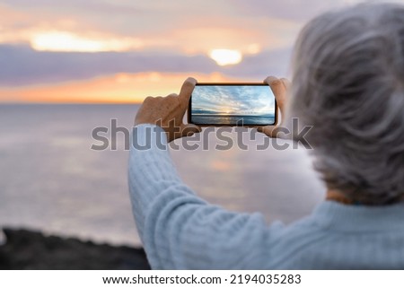 Woman holding her mobile phone taking a picture of the sunset over the sea, dramatic sky and sunlight