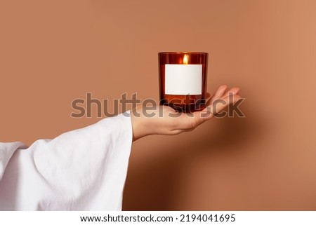 Covered with a white cloth hand holding a candle on a brown background. Mockup for candle shop. Aromatherapy poster. Space for the logo. Boho atmosphere for scent products. AD banner for the spa. 