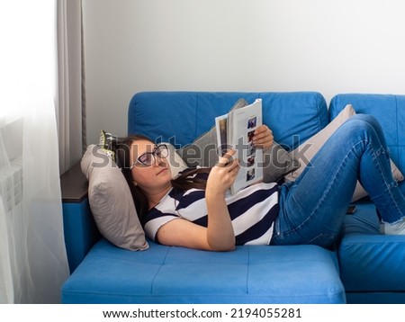 Woman with dark hair in a T-shirt lying on a blue sofa in her living room and reading the news. Female in glasses at home reading article in magazine. Studying at home.