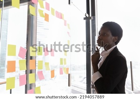 Thoughtful serious African American businesswoman thinking over project tasks management. Startup manager looking at sticker notes on glass board, analyzing strategy, plan of work Royalty-Free Stock Photo #2194057889