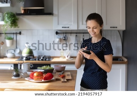 Cheerful chef blogger woman using mobile phone at kitchen table, preparing salad from fresh vegetables, healthy food ingredients, browsing online recipe on Internet on smartphone