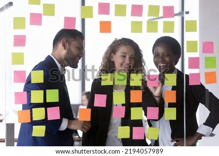 Happy multiethnic business team writing tasks on colorful notes, meeting at glass board, working on project plan, strategy, spreading tasks, talking, smiling, laughing, studying scrum management Royalty-Free Stock Photo #2194057989