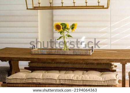 Modern Stylish Interior Design in Dining Room with Table Centered Under Chandelier and Bouquet of Sunflowers as Centerpiece. Table is Flanked by Benches with Cushions. Background is Automated Electric Royalty-Free Stock Photo #2194064363