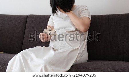 Photo of a pregnant woman covering her face while sitting on a sofa and holding a house in her hand