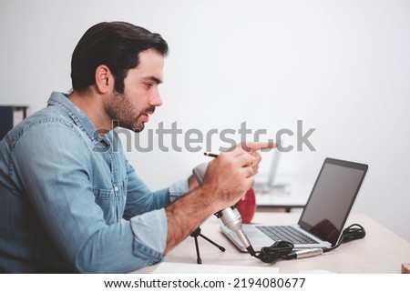Young man host sitting at table, streaming audio podcast using microphone and laptop in studio, Social media, podcasting, blogging concept. Cheerful casual caucasian employee.