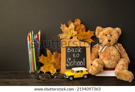 On a black background, a soft teddy bear, a book, glasses, a yellow typewriter, a school board with text, autumn leaves, colored pencils sit on the table.  Back to school concept, copy space for text.