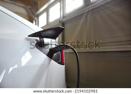 White EV electric car plugged with battery charger and charging in garage at home. At home charing station brings more convenient, less expensive and more cost savings comparing to supercharger. Royalty-Free Stock Photo #2194102981