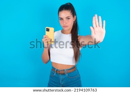 beautiful brunette woman wearing withe tank top shirt over blue background  using and texting with smartphone with open hand doing stop sign with serious and confident expression, defense gesture