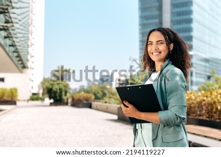 Successful positive confident young brazilian or hispanic curly haired business woman, real estate agent, manager, holds documents, stand outdoors near business center, looks at camera, smile Royalty-Free Stock Photo #2194119227