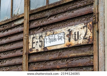 An old rusty lamella door with a sign that says "E-Spulen 1+2" (E-coils 1+2) in German.