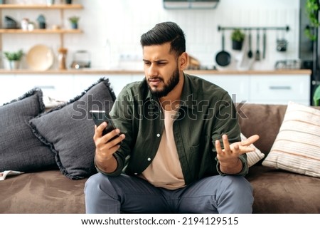 Disappointment puzzled mixed race young man in casual wear, sits on sofa in living room, using mobile phone, texting online, browsing social networks, reads news, looks at screen in confusion Royalty-Free Stock Photo #2194129515