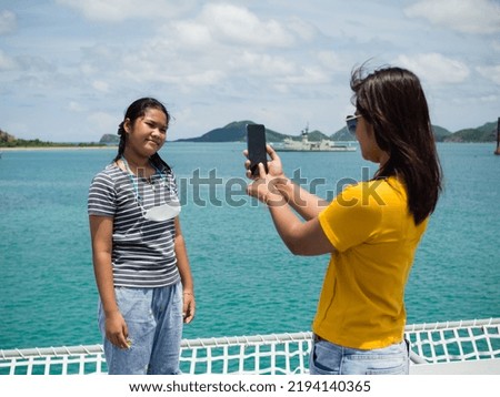 A woman in a yellow shirt is holding a phone to take pictures of a girl in a gray shirt. With a background of blue water and small mountains. It is tourism after the corona virus outbreak.