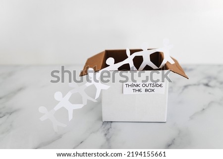 Think outside the box concept with paper people chain getting out of a white box with text on white marble	with minimalistic composition