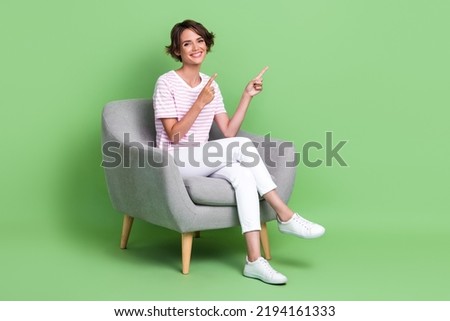 Full length photo of pretty young girl sit chair fingers point empty space wear stylish striped garment isolated on green color background