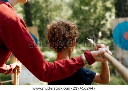 Back view of little curly boy learning to throw a javelin at a wooden target, outdoors. Festival of medieval culture and games. Royalty-Free Stock Photo #2194162447