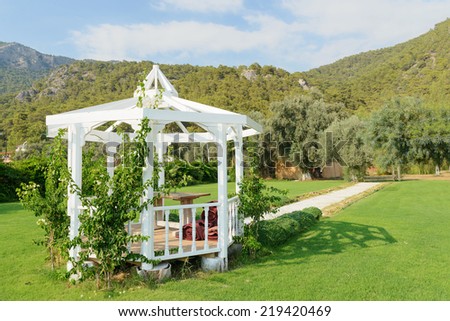 Image of a garden pergola on a sunny day with mountains and forests in the background against a blue sky with white clouds