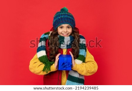 smiling teen girl wearing knitted hat hold present box on red background, xmas