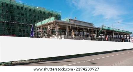 Long blank hoarding with white canvas mounted on construction site with unfinished building with wall and scaffold