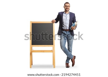 Full length portrait of a male teacher holding books and standing next to a blackboard on a stand isolated on white background