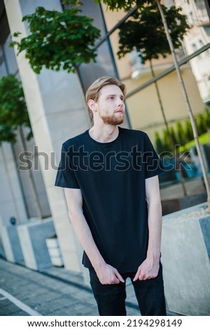 a young hipster in a black t-shirt stands on the street. Place for a logo