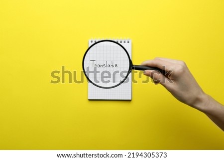 Woman holding magnifying glass over sheet of paper with word Translate on yellow background, top view