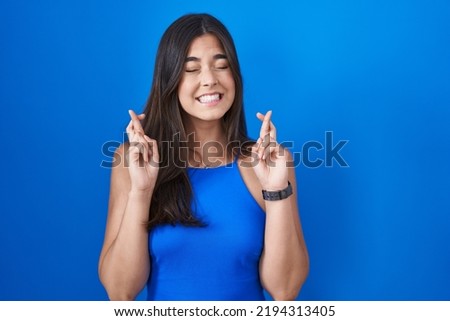 Hispanic woman standing over blue background gesturing finger crossed smiling with hope and eyes closed. luck and superstitious concept. 