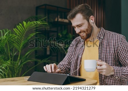 Young fun man he wear shirt earphones work study on tablet pc computer drink tea sit alone at table in coffee shop cafe relax rest in restaurant in free time. Freelance mobile office business concept