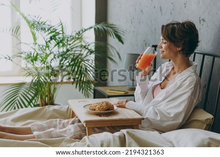 Side view young woman wear white shirt pajama she lying in bed eat breakfast drink orange juice rest relax spend time in bedroom lounge home in own room hotel wake up dream be lost in good mood day. Royalty-Free Stock Photo #2194321363
