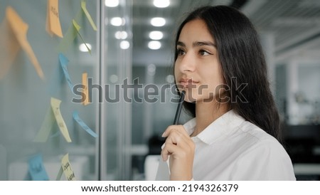 Arabian business lady managing daily tasks stand near board in office write reminder information instruction on paper sticky notes read stickers create to-do list analyze work organize corporate