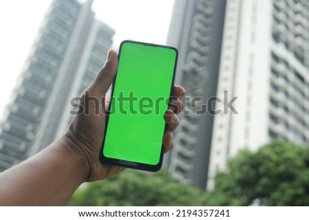  young man holding smart phone with green screen against city buildings 