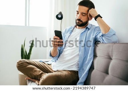 Young brazilian man sitting on sofa using smartphone