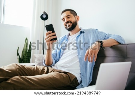 Smiling man sitting on couch at home using laptop and cell phone Royalty-Free Stock Photo #2194389749