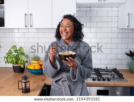 A young multi-ethnic woman laughs while eating breakfast cereal in dressing gown Royalty-Free Stock Photo #2194393181