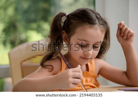 Close-up portrait of a beautiful European little girl of preschool age with two ponytails, in an orange top, sitting at a table with a paintbrush in her hand and painting with watercolors