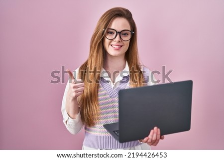 Young caucasian woman working using computer laptop doing happy thumbs up gesture with hand. approving expression looking at the camera showing success. 