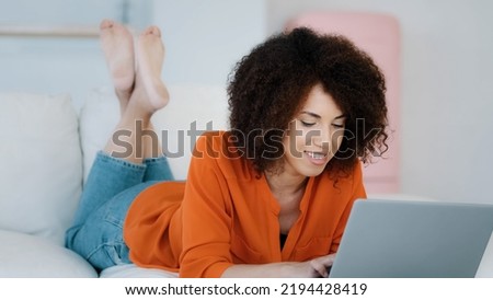 African 20s lady 30s woman freelancer with curly hair lying on sofa relaxing typing on laptop making online purchase in internet store app working remotely from home chatting with friends with