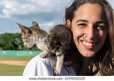 Close-up portrait of a smiling young Latin girl outdoors with a kitten on her shoulder, she has a small silver piercing in her nose