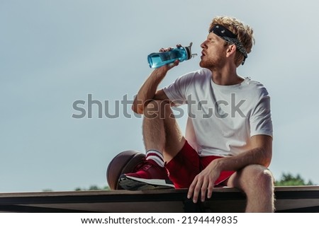 sportsman in white t-shirt drinking from sports bottle while sitting near ball