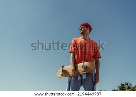 low angle view of man in sunglasses and beanie standing with skate under blue sky