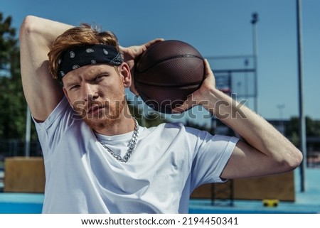 redhead man in bandana and necklace holding ball and looking at camera