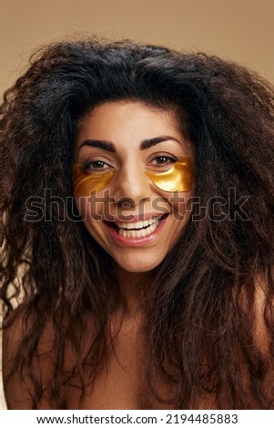 Happy curly pretty Latin female with gold hydrogel patches under eye laugh at camera posing isolated over pastel beige background. Cosmetic product ad Natural beauty concept Studio portrait