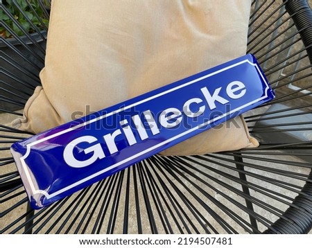 Close up of Street Sign Grillecke BBQ Corner Blue and white on black wired Garden chair and beige pillow