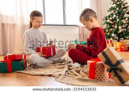 christmas, winter holidays and childhood concept - happy girl and boy in pajamas opening gifts sitting on floor in front of each other at home