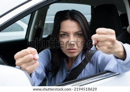 A very angry aggressive woman is clenching her fists and threatening from her  car. Royalty-Free Stock Photo #2194518629