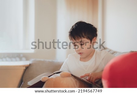 Happy School kid sitting on sofa reading a book with morning light shining from window. Child boy reading story relaxing at home on weekend, Positive children concept