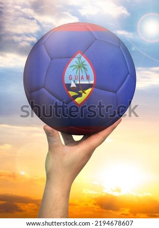 Soccer ball in hand with a depiction of the flag of Guam against a colorful sky