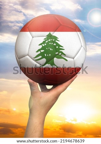 Soccer ball in hand with a depiction of the flag of Lebanon against a colorful sky