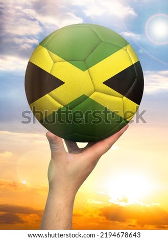 Soccer ball in hand with a depiction of the flag of Jamaica against a colorful sky