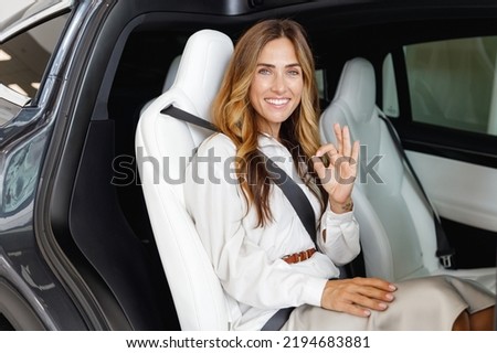 Happy woman customer buyer client in white shirt using safety belt show ok gesture choose auto want buy new automobile in car showroom vehicle salon dealership store motor show indoor. Sales concept. Royalty-Free Stock Photo #2194683881