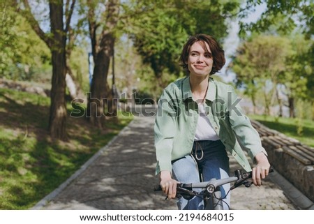 Young pensive dreamful happy woman 20s wearing casual green jacket jeans riding bicycle bike on sidewalk in city spring park outdoors, look aside. People active urban healthy lifestyle cycling concept Royalty-Free Stock Photo #2194684015
